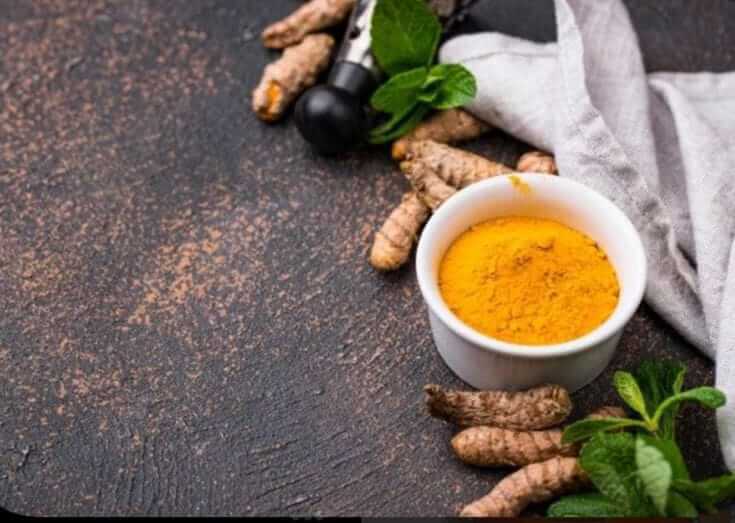 Jamu Organic Herbal with turmeric and ginger roots in a white bowl, surrounded by fresh herbs, on a dark textured surface.